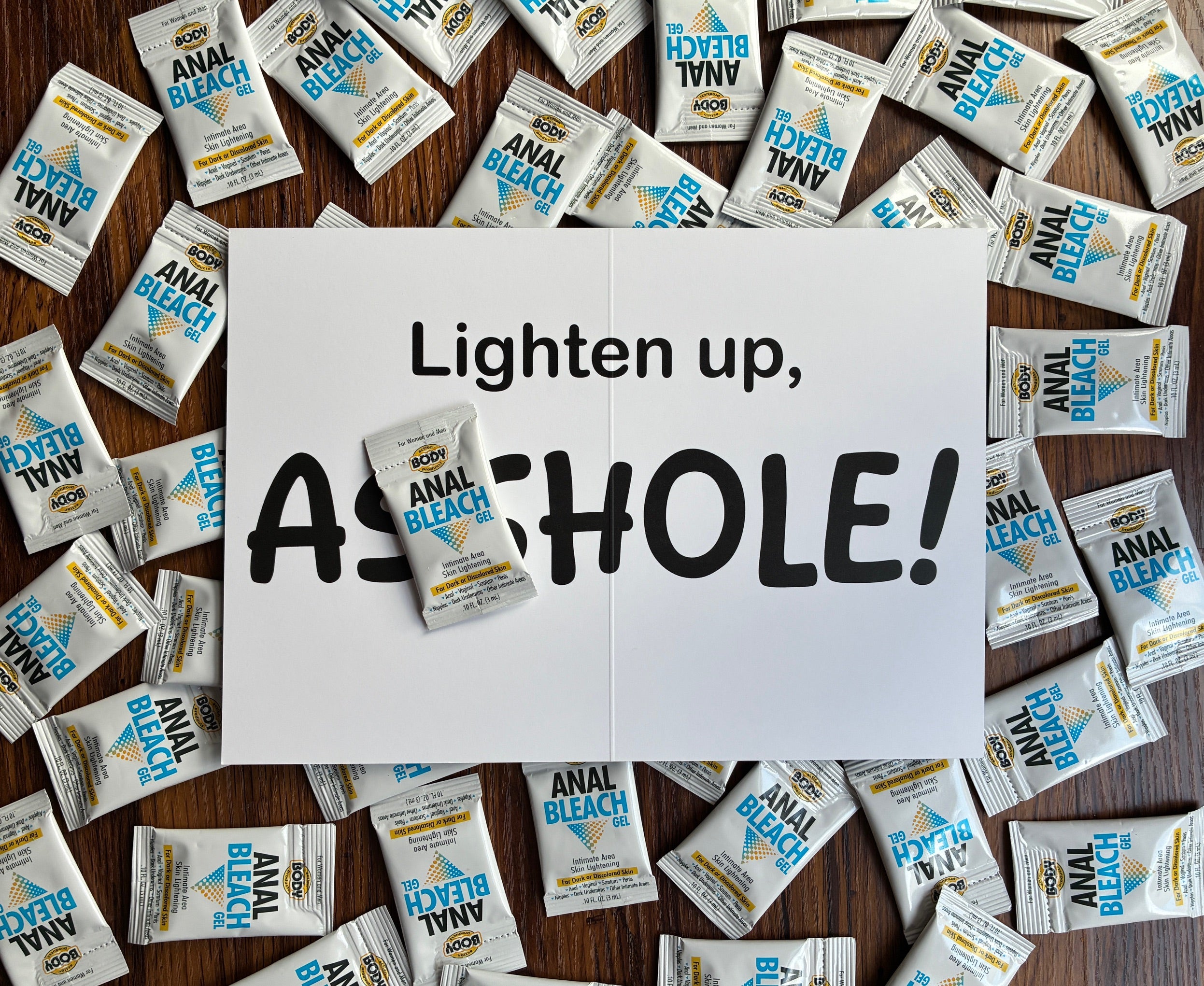 Interior of "Lighten up Asshole" prank card with bold text and sample anal bleach packet on a wooden surface, surrounded by additional anal bleach packets